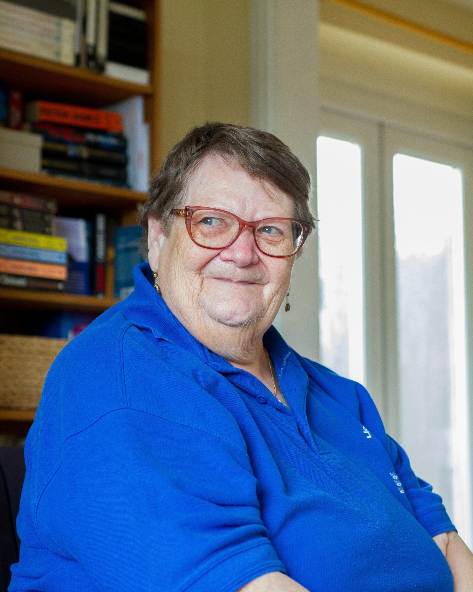 An older woman in a blue shirt and red glasses, smiling, in a room lined with books