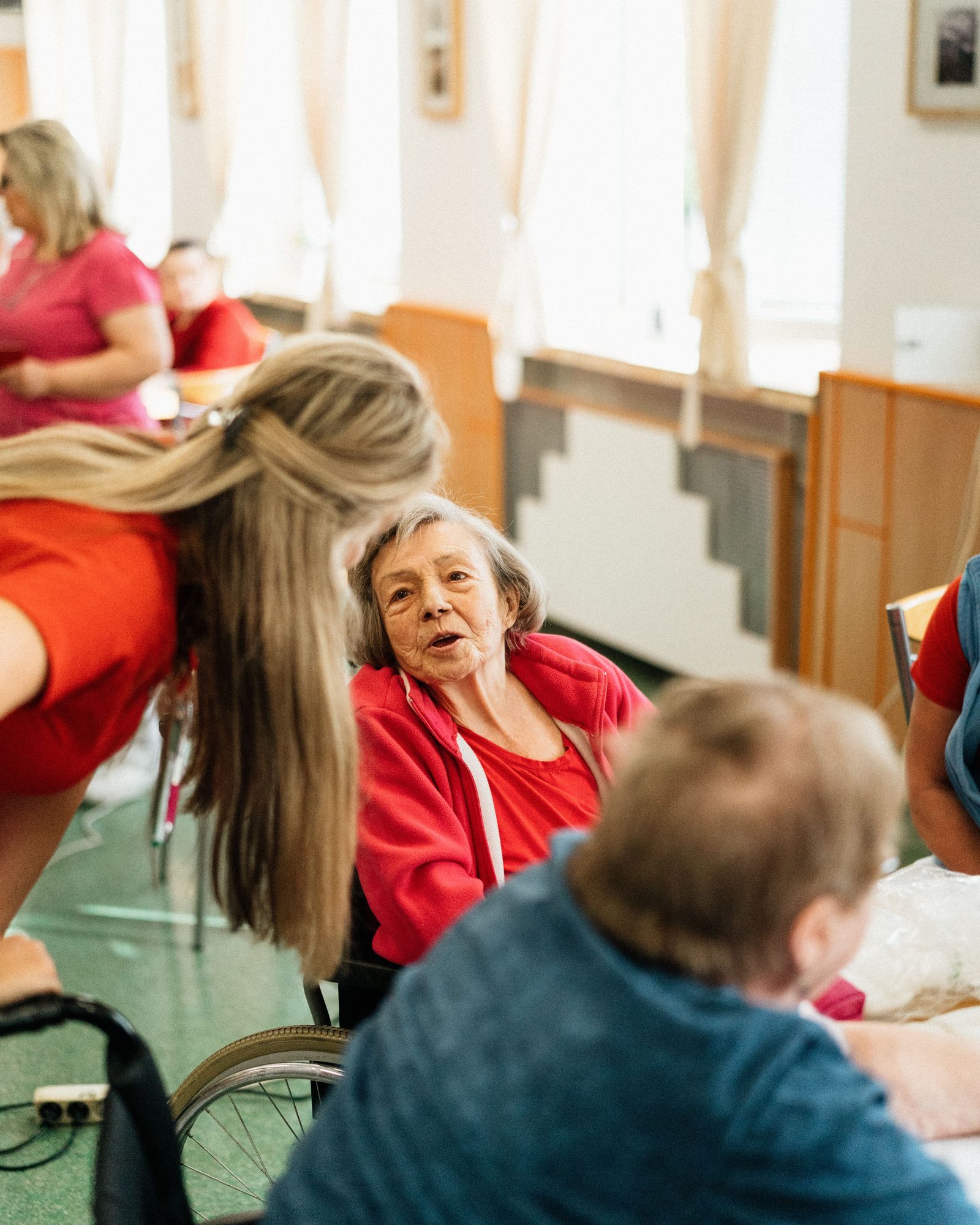 An older woman in a red cardigan talking with a caregiver in a bright common room